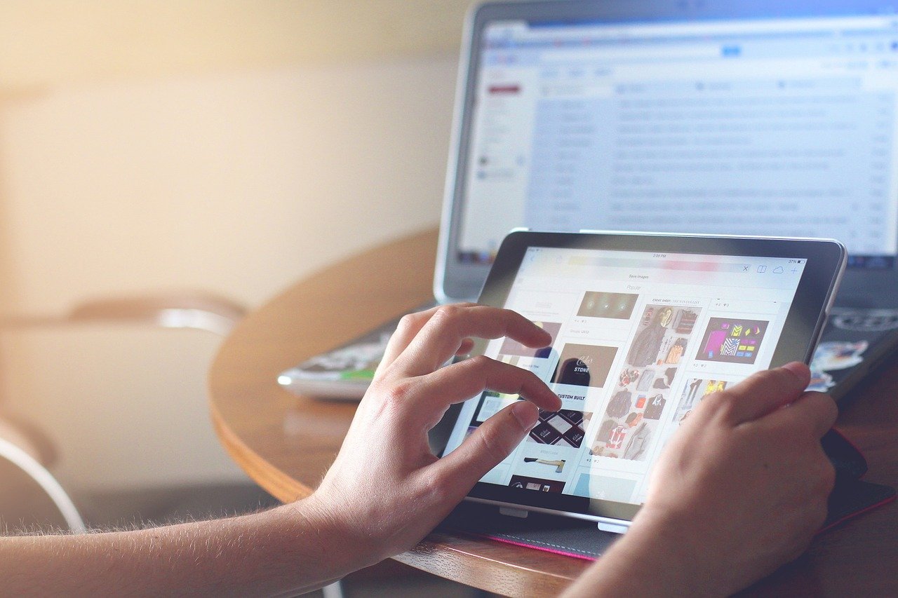 A person sitting at a table. They are holding a touch screen tablet and there is a laptop sitting on the table behind the tablet. 