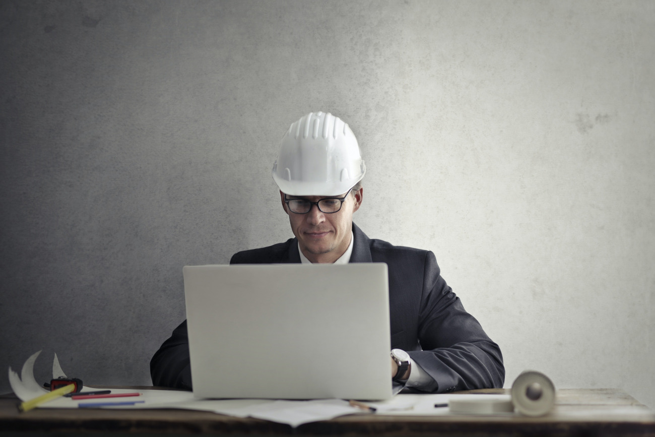 A man facing the camera. He is sitting at a desk using a laptop computer. He's wearing a nice business suit--and oddly, a hard hat.
